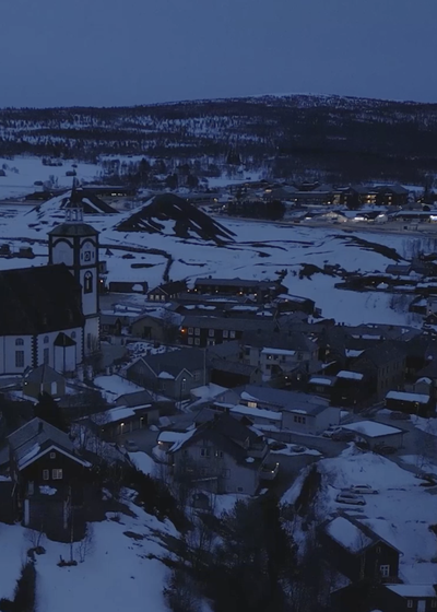 Røros sentrum fra droneperspektiv i blåtimen med Røros Kirke som fokus og opplyst trehusbebyggelse rundt.