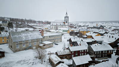 Dronebilde som viser fasaden til Uldvarefabrikken med Røros kirke i bakgrunnen og snødekt trehusbebyggelse rundt fasaden.